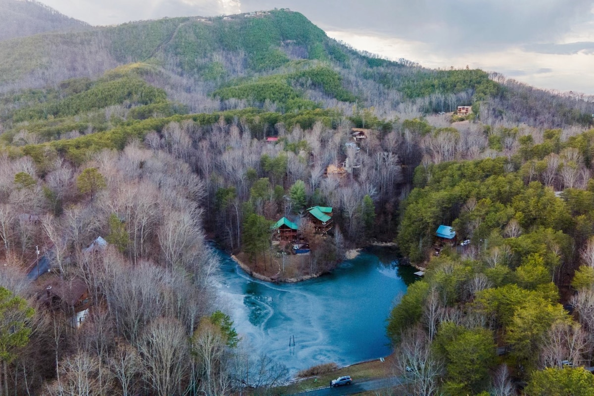 A tranquil aerial view captures the cabin nestled among a dense forest, surrounding a serene lake. Bare trees reflect the transition of seasons, while a hint of water glistens in the foreground, complemented by the distant mountain slopes under a cloudy sky.