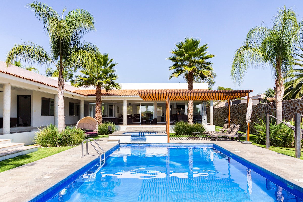 A tranquil outdoor space is highlighted by a crystal-clear pool surrounded by lush palm trees. A covered terrace provides shade and seating, with the home visible in the background. The well-maintained grounds contribute to a serene atmosphere.
