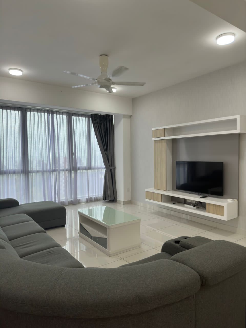 A spacious living area features a curved grey sofa and a modern coffee table. A wall-mounted television is placed on a light wooden media unit. Large windows are covered with sheer curtains, allowing for ample natural light.