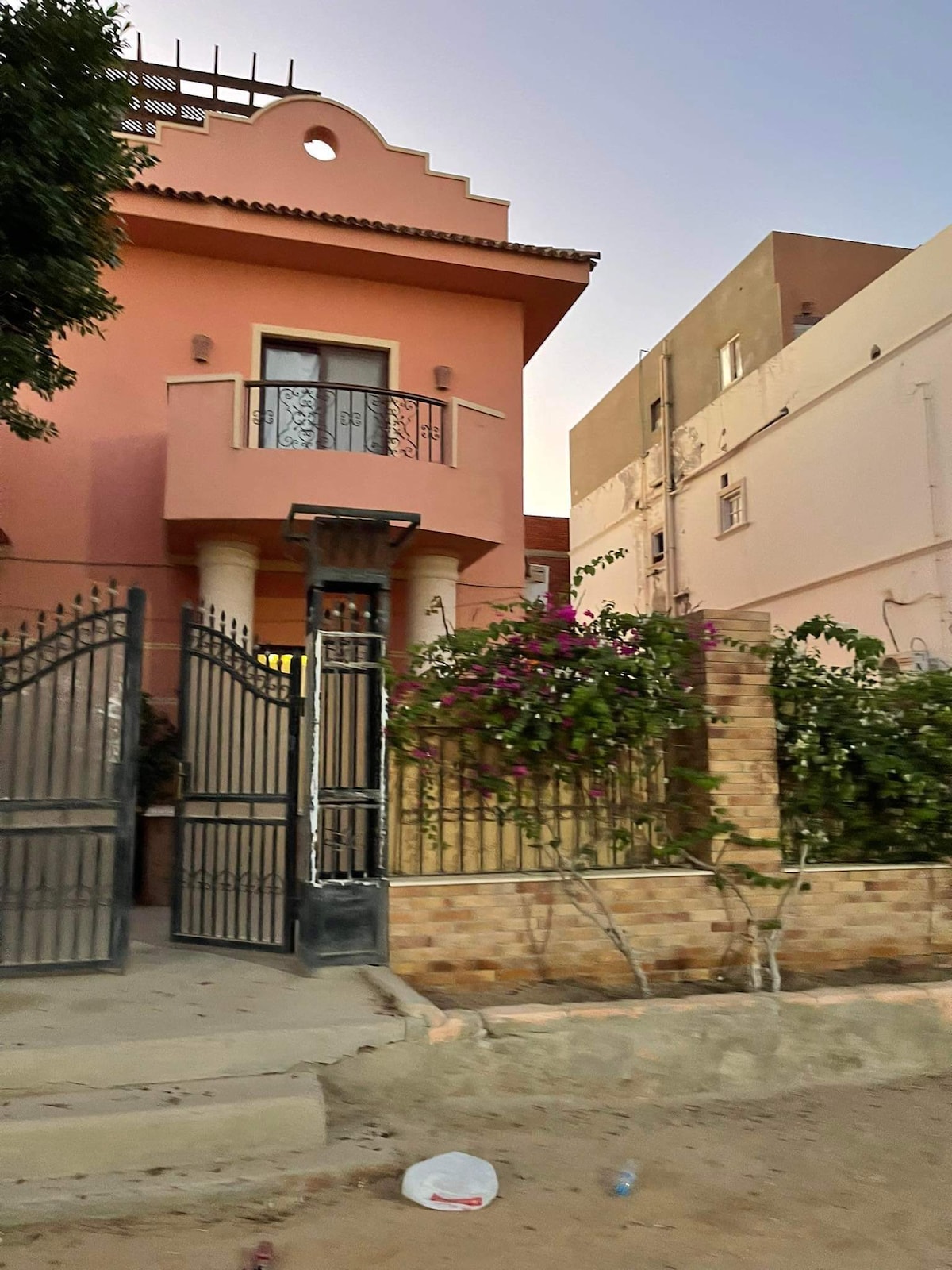 A large villa is enclosed by a wrought iron gate, showcasing vibrant bougainvillea climbing along the stone wall. The exterior features warm-toned stucco and a balcony with decorative railings above the entrance, providing a welcoming approach to the property.