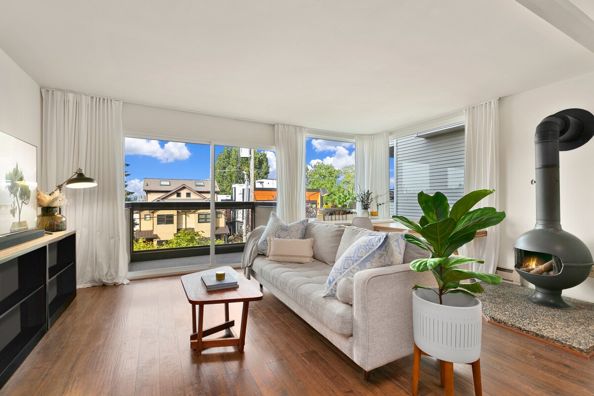 A spacious living area features large glass doors that open to a view of greenery and nearby homes. A cozy sofa is positioned beside a small coffee table, while a unique fireplace adds charm to the space. Light drapes soften the natural light filtering in.