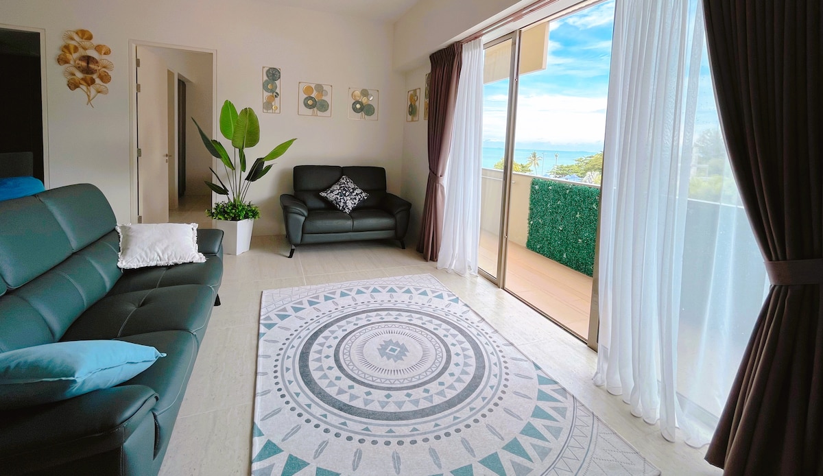 A bright living area features a modern design with large glass doors opening to a scenic view. A patterned rug lies on the floor, complemented by two dark sofas and a decorative plant. Natural light enhances the open space, creating a serene ambiance.