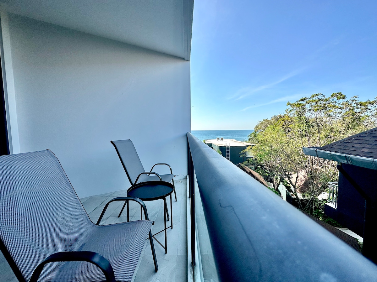 A balcony is shown with two grey lounge chairs positioned beside a glass railing. In the distance, an unobstructed view of the ocean is visible, complemented by a clear blue sky. Lush greenery is partially seen on one side of the image.