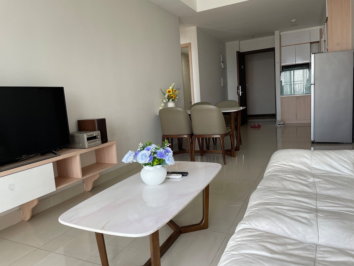 A light-filled living space features a white leather sofa and a modern, low coffee table. A vase with blue flowers sits on the table, while a television stands on a wooden stand. A dining area with four beige chairs is visible in the background.