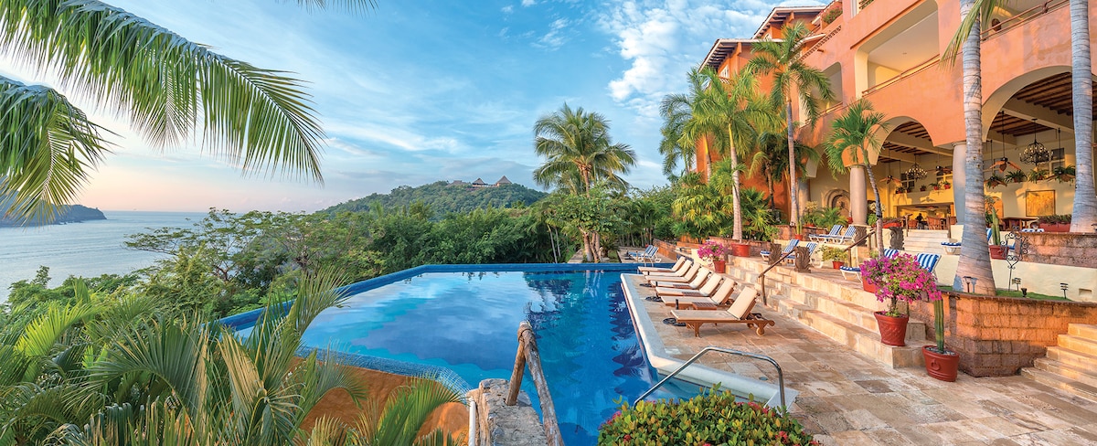 An outdoor pool is framed by lush tropical gardens and loungers under tall palm trees. The clear water reflects the sky and surrounding hills, while an outdoor dining area embraces the view. The resort building blends harmoniously with the natural landscape.