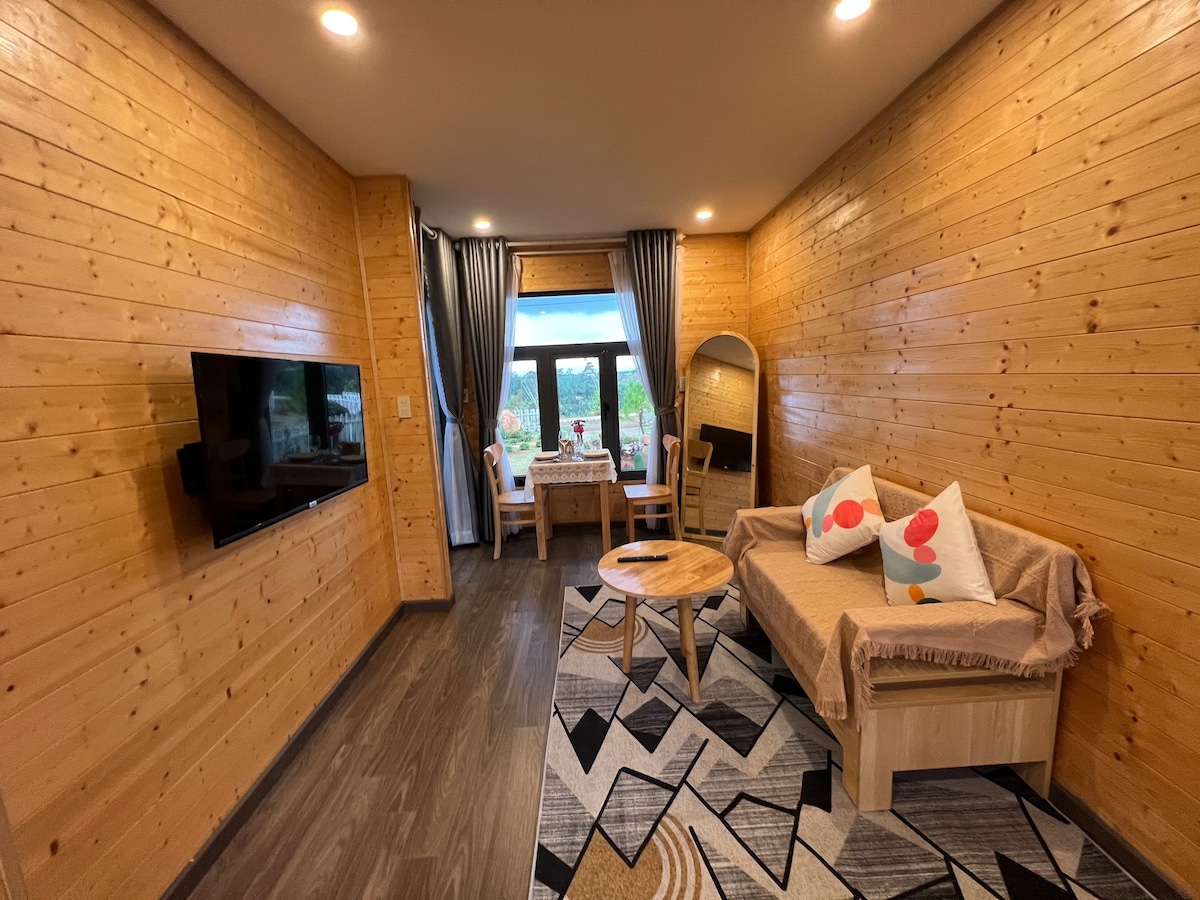 A wooden paneled living space is equipped with a comfortable seating area. A small table is positioned in front of a sofa adorned with colorful cushions. Large windows invite natural light and offer outdoor views, while soft drapes frame the windows.