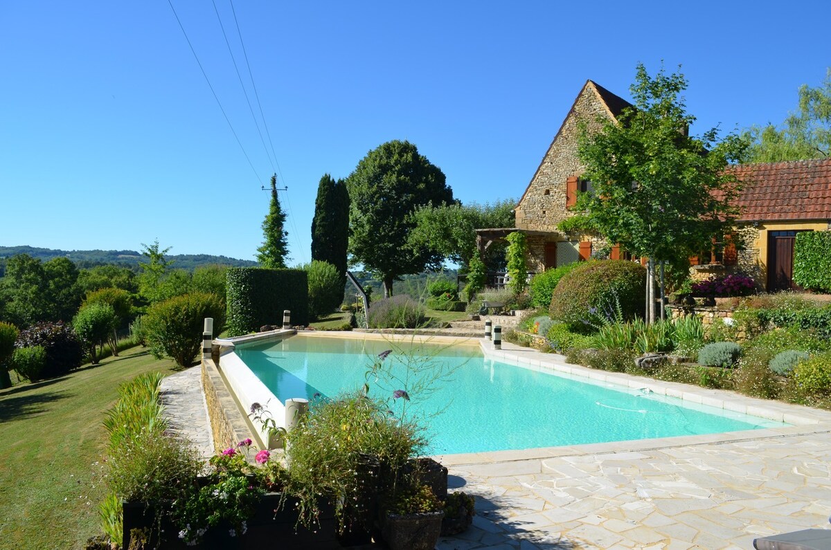 A spacious outdoor swimming pool is framed by lush gardens and neatly trimmed hedges. The stone structure of the house is visible in the background, complemented by trees and shrubs. Clear blue skies and well-maintained paths enhance the serene outdoor setting.