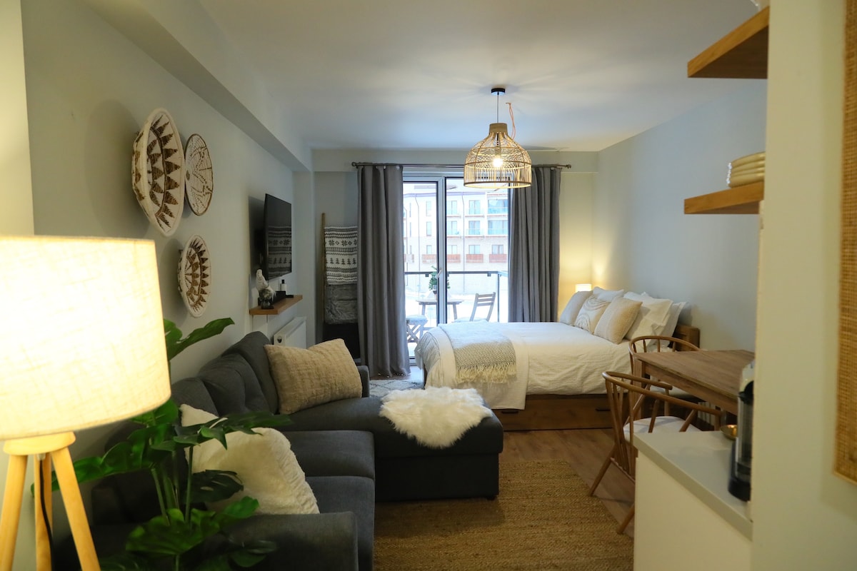 A studio apartment is shown, featuring a spacious bed adorned with light bedding. A cozy seating area includes a sectional couch and a small dining table. Natural light enters through a glass door leading to a balcony, while modern decor elements are displayed on the walls.