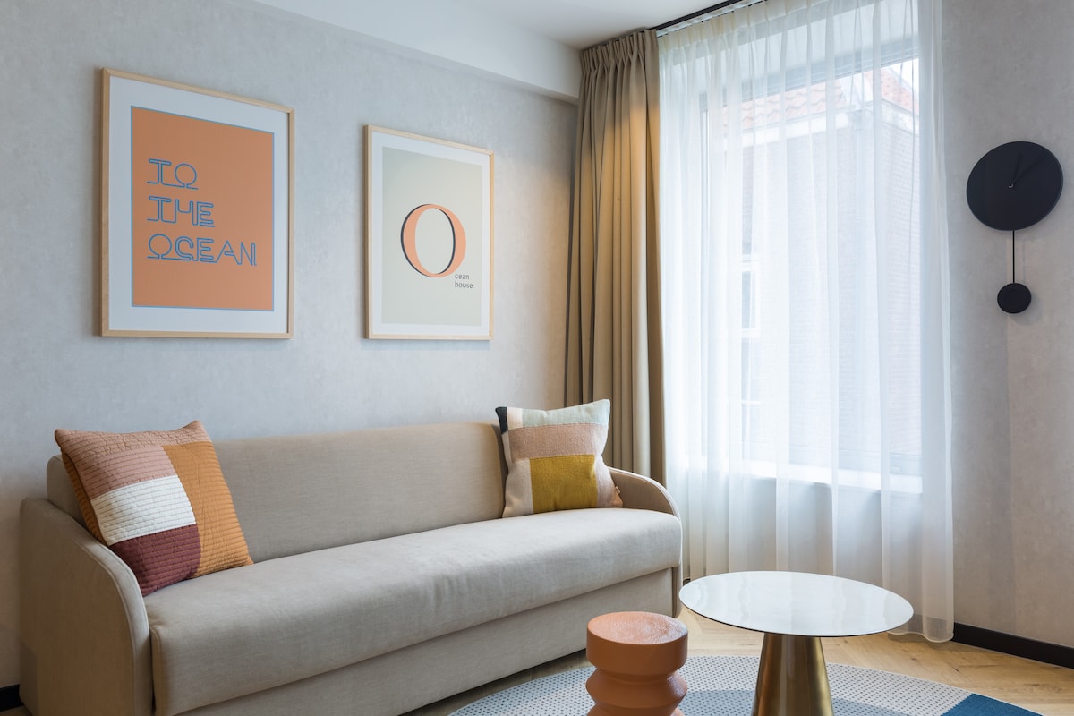A light-filled living area features a comfortable sofa adorned with decorative cushions. Art prints are displayed on the wall, and a round coffee table sits on a textured rug. Sheer curtains allow natural light to softly illuminate the space.