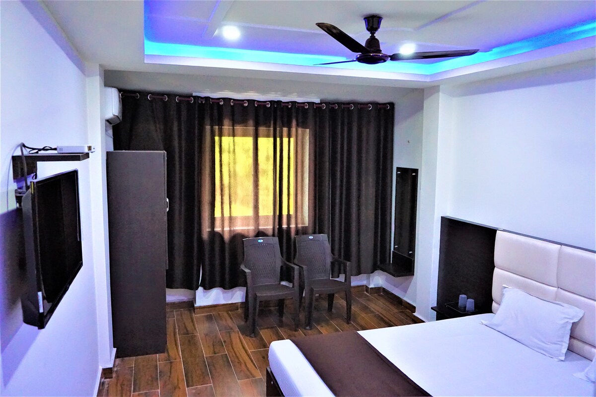 A well-lit guest room is furnished with a spacious bed and two dark chairs positioned near a window. A flat-screen television is mounted on the wall, alongside a dark wardrobe and a light-colored bedside panel. Dark curtains frame the window, allowing for privacy.