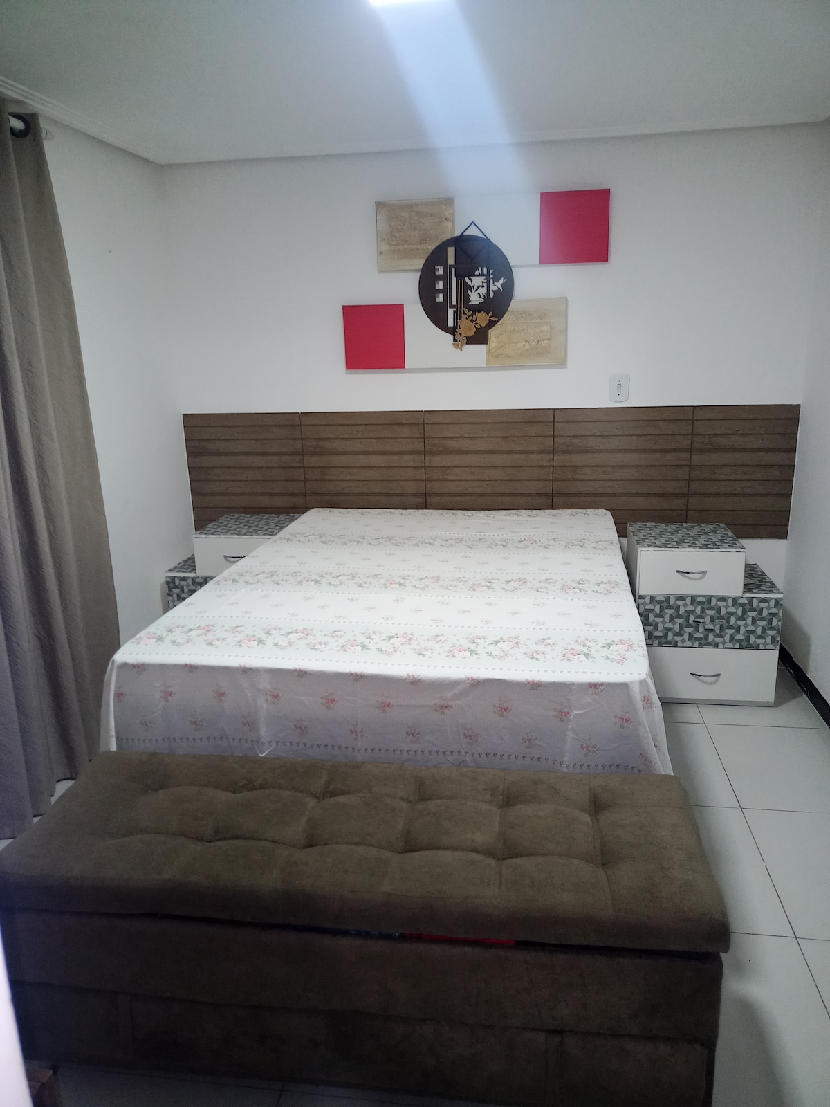 A sleeping area features a spacious bed with a floral-patterned coverlet against a wooden headboard. Flanking the bed are two bedside tables, and a cozy bench is positioned at the foot of the bed. Soft light brightens the room, creating a serene ambiance.