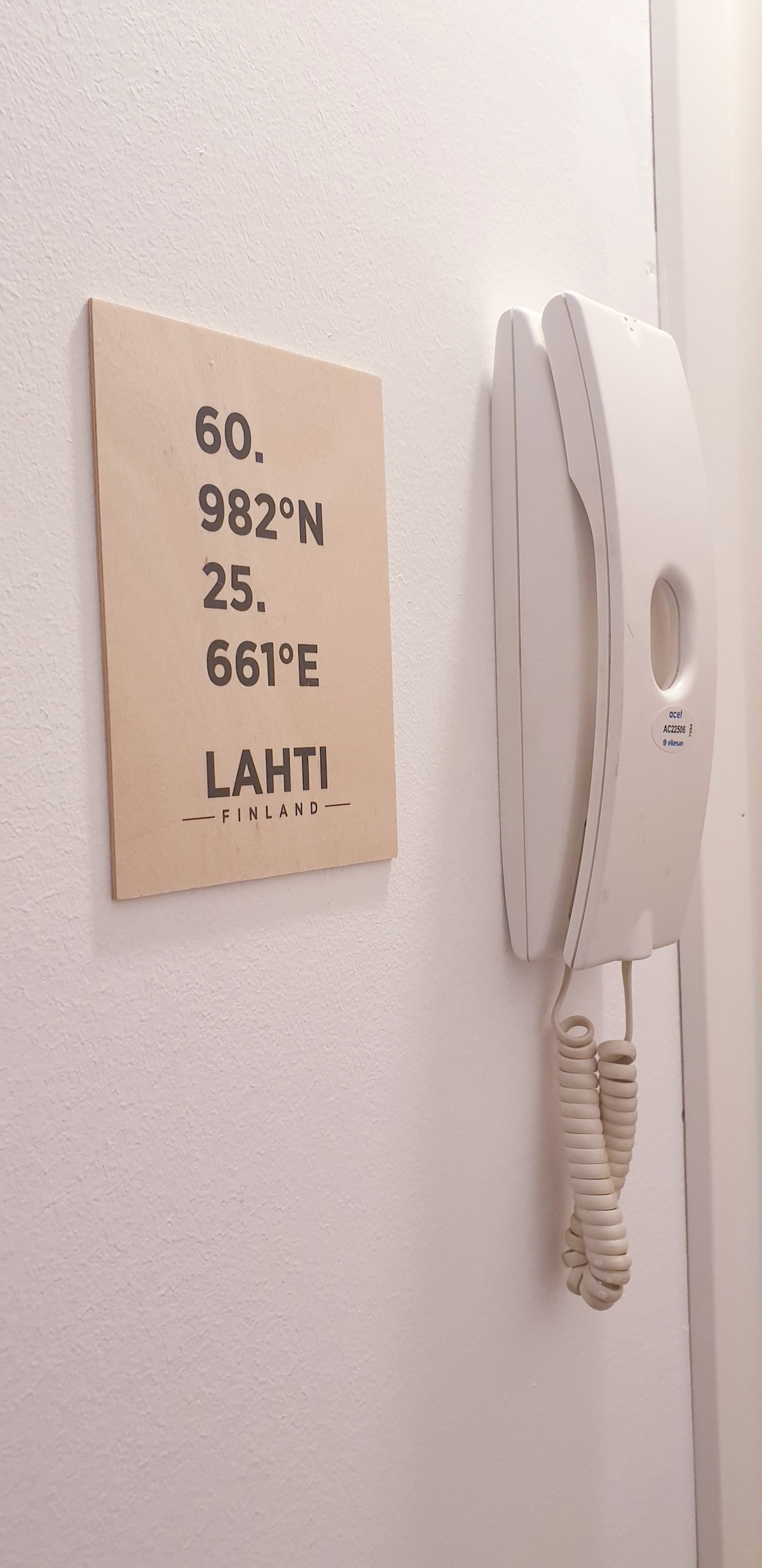 A wooden sign displays the geographical coordinates of Lahti, Finland, accompanied by a wall-mounted telephone. The sign features a simple design, while the telephone is positioned beside it, presenting a functional aspect of the room.