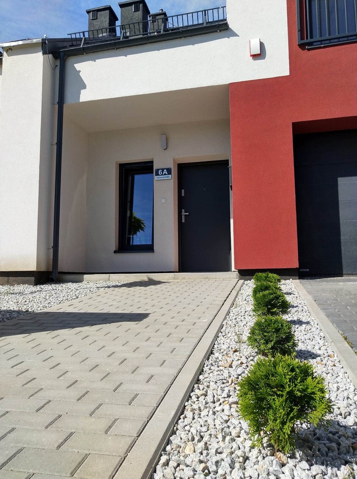 The entrance to the building is framed by a neatly paved pathway, bordered by decorative gravel and small, manicured shrubs. A modern door, tinted windows, and a building facade featuring contrasting colors create an inviting appearance.