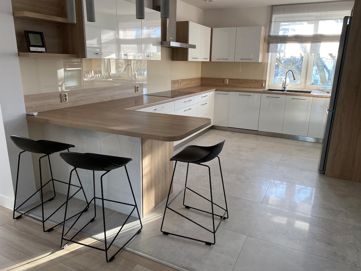 A modern kitchen with sleek cabinetry and a large island is visible. Two high black stools are positioned at the island, and ample natural light streams in through a window above the sink, illuminating the neutral tile flooring.