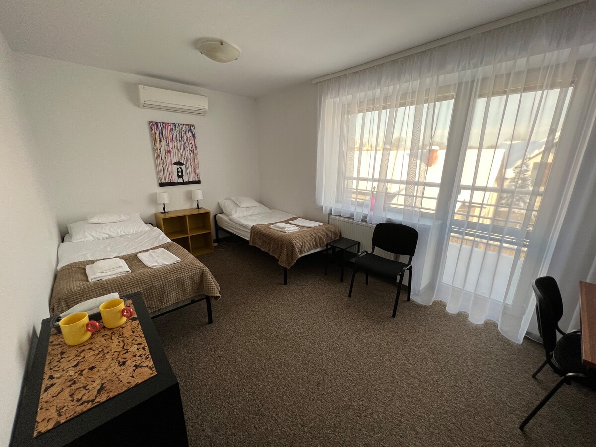 A well-lit bedroom includes two single beds and one larger bed, arranged with simple bedding. A wooden chair and a desk occupy one corner. Full-length curtains adorn a large window, allowing natural light to fill the space. A decorative artwork is displayed on the wall.