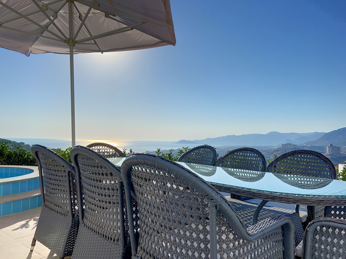 A dining area is set with a large glass table surrounded by comfortable woven chairs, shaded by an umbrella. The backdrop features a stunning view of the sea and distant hills under a clear blue sky.