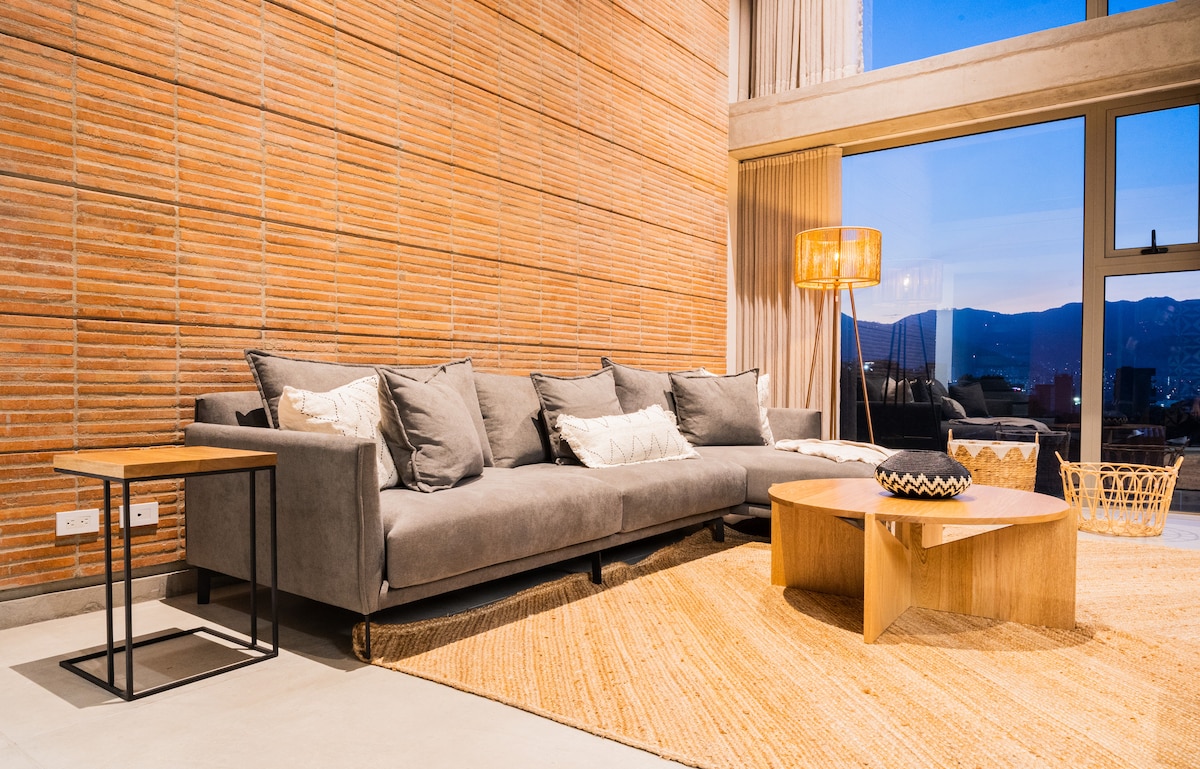 A spacious living area features a large gray sectional sofa adorned with decorative cushions. A wooden coffee table sits at the center, alongside a small side table. Natural light enters through large windows, illuminating the textured brick wall and adding warmth to the space.