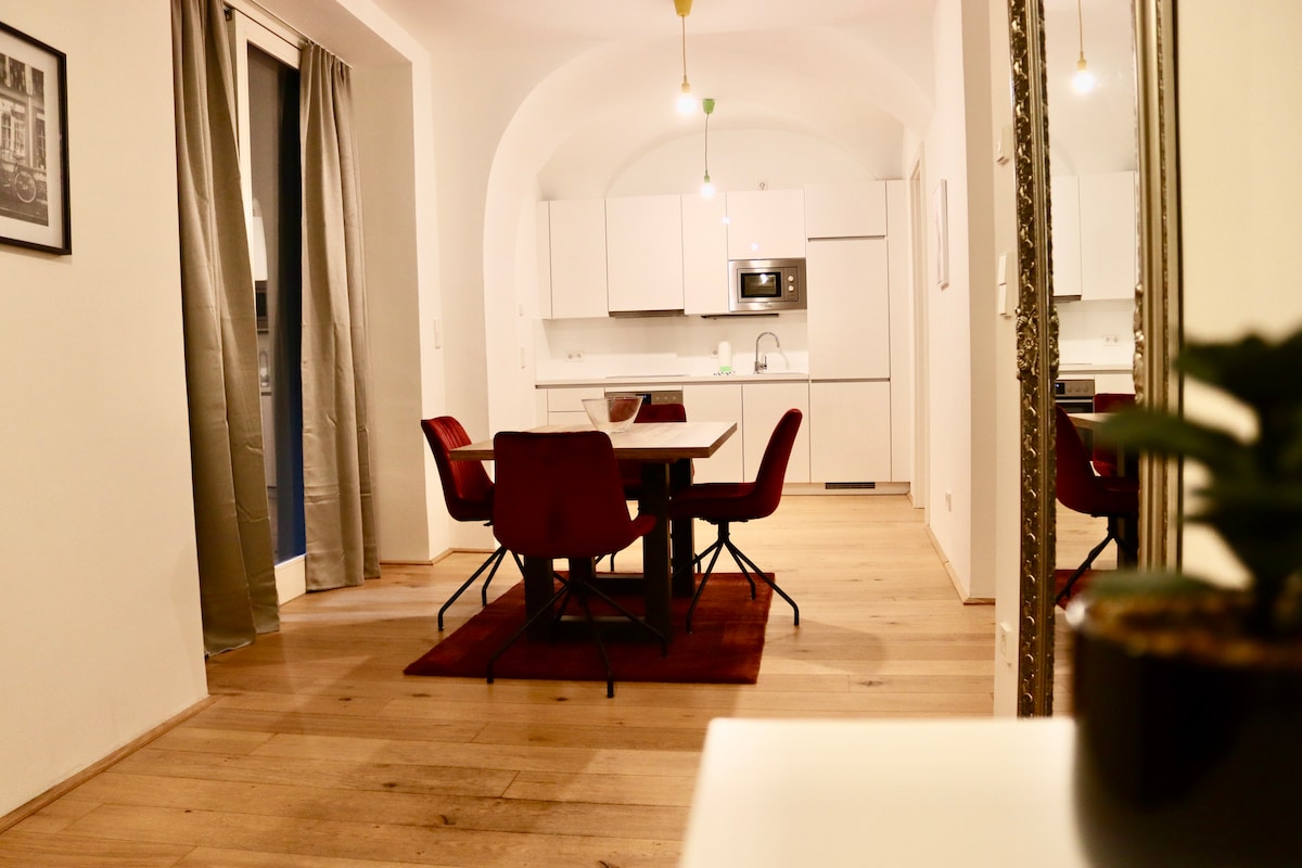 A modern dining area is presented with a round table surrounded by four red chairs. The kitchen is visible in the background, featuring sleek white cabinetry and stainless-steel appliances. Natural light enters through large glass doors, highlighting the warm wooden floor and neutral-toned walls.