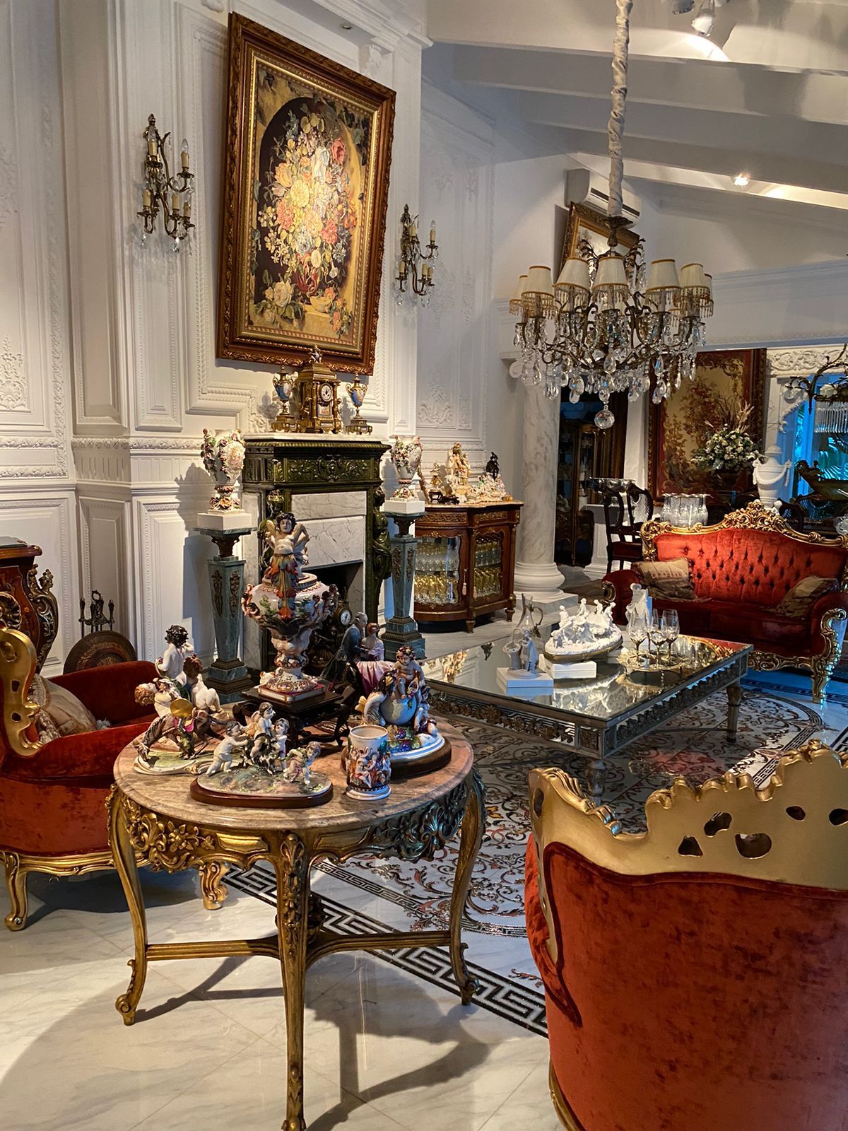 An elegantly decorated living area features opulent red chairs with golden accents, a detailed wooden table displaying ornamental figurines, and a classic chandelier above. The walls are adorned with refined artwork, while a stylish rug adds warmth to the marble floor.