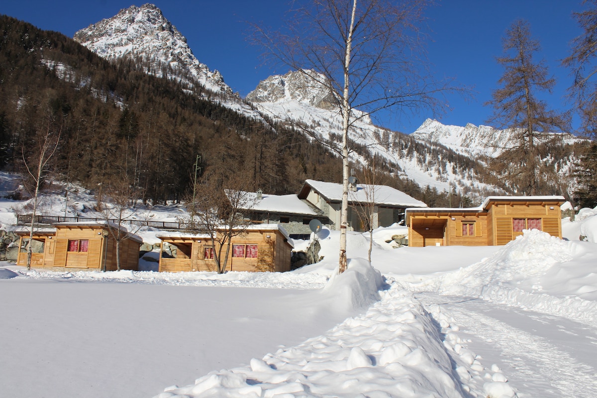 The image captures a cluster of wooden cabins set against a backdrop of snow-covered mountains. A path lined with snow leads towards the cabins, highlighting the serene winter landscape, while patches of clear sky provide a contrast to the white surroundings.