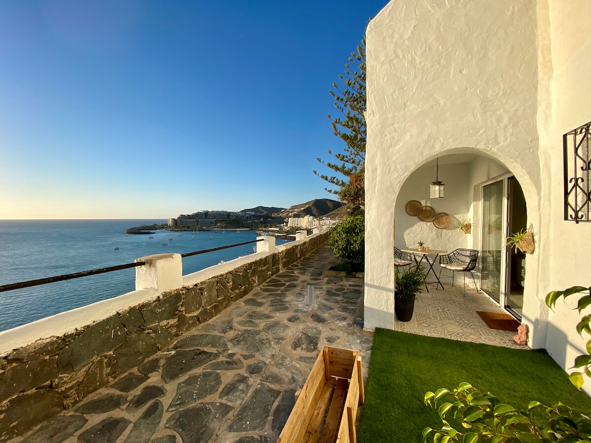 A charming terrace overlooks the sea with a pathway made of rustic stone. A small round table and chairs are positioned on the terrace, adorned with decorative wall accents. The coastal view extends along the horizon, capturing the serene water and distant beach.