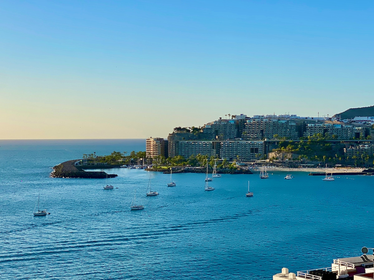 A tranquil view of the coastline in Gran Canaria is depicted, showcasing calm waters with several sailboats gently drifting. The shoreline is lined with palm trees and buildings, set against a clear blue sky as the sun begins to set, casting a warm glow over the scene.