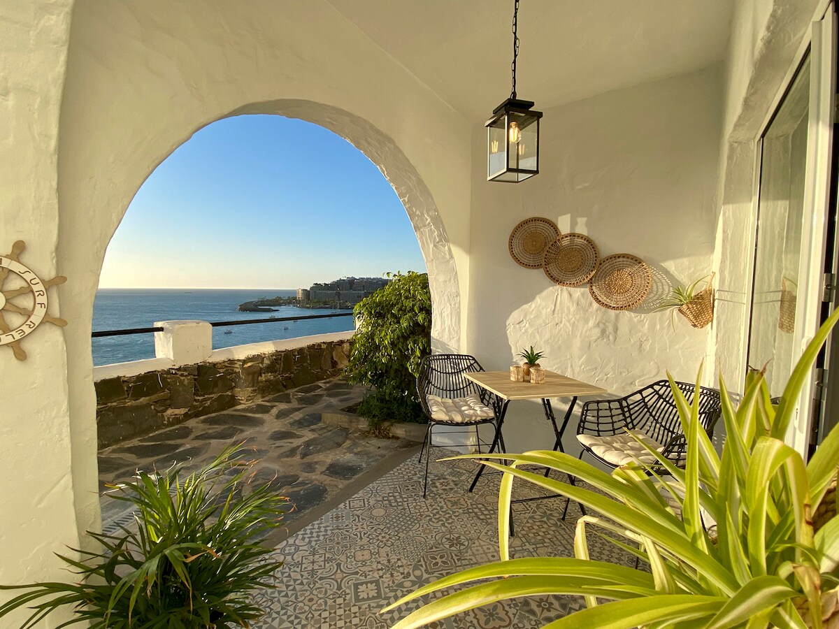 A terrace is framed by an arched opening, featuring a small table and two chairs. The scenic view includes the ocean and distant coastline. Decorative elements, such as woven wall art and potted plants, add to the cozy ambiance.