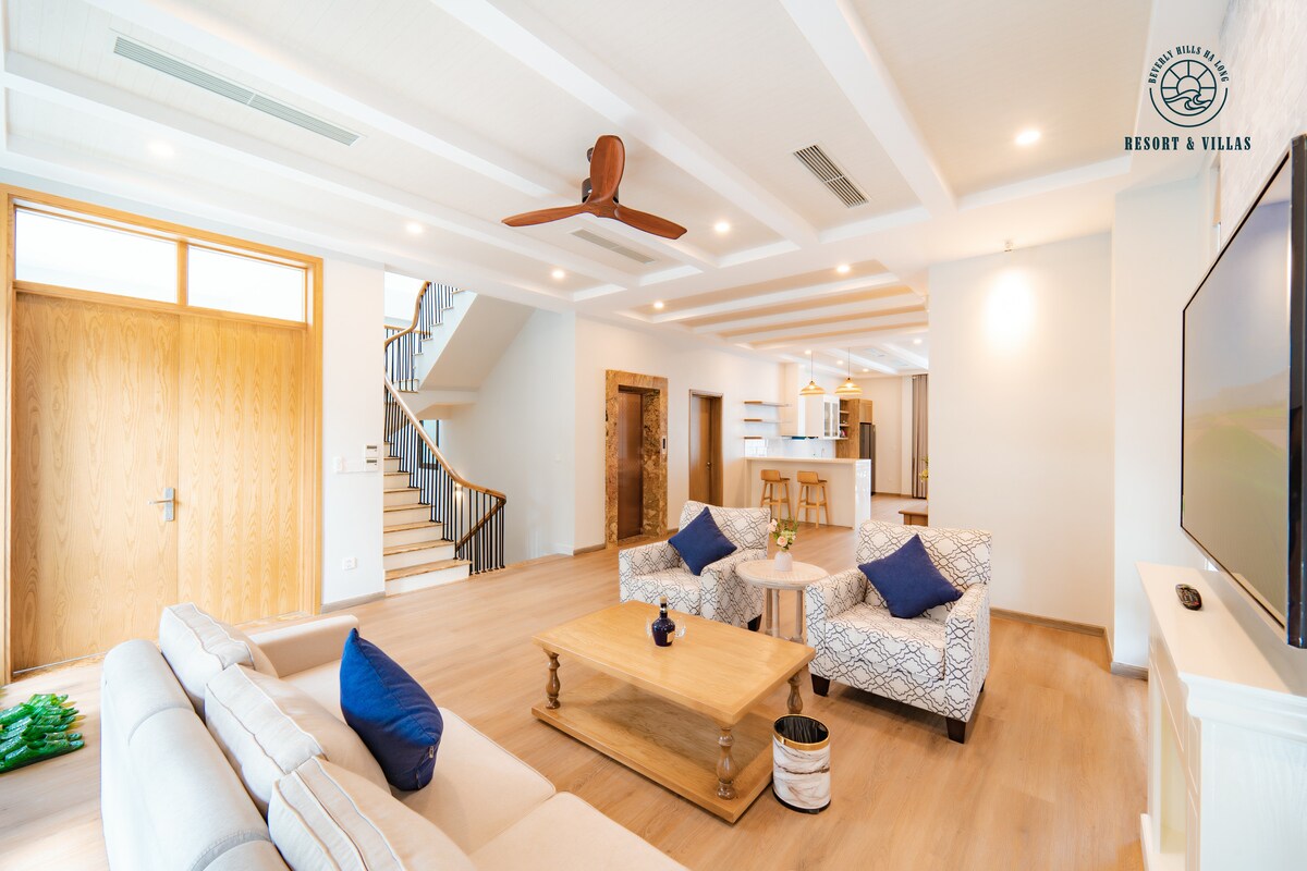 A bright and open living space features light-colored furniture arranged around a wooden coffee table. Decorative blue pillows accent the sofas. Large windows allow ample natural light, and a staircase is visible in the background, leading to an upper level.