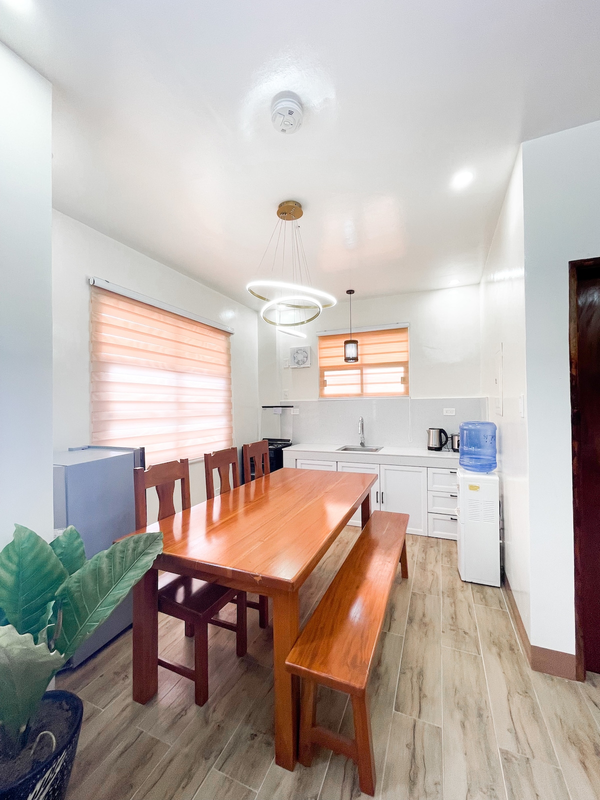 A modern kitchen area features a wooden dining table accompanied by bench seating. Large windows with light-filtering shades allow natural light to fill the space. Appliances, including a refrigerator and a water dispenser, are visible, complemented by minimalist cabinetry and a stylish overhead light fixture.