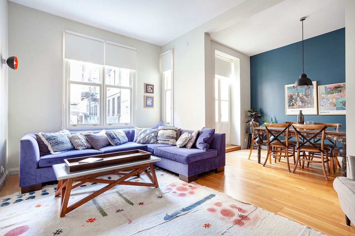 A spacious living area features a deep blue sofa adorned with decorative pillows. Natural light filters through large windows, illuminating the hardwood floor. A dining table with wooden chairs is positioned against a navy accent wall, complemented by vibrant artwork and greenery in the corner.