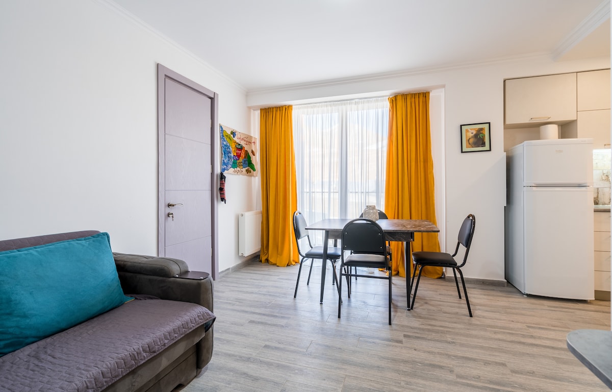 A light-filled living area features a gray sofa adorned with a turquoise cushion, accompanied by a dining table set for four. Bright yellow curtains frame a large window, complementing the modern kitchen area with a refrigerator. A colorful artwork adds visual interest to the space.