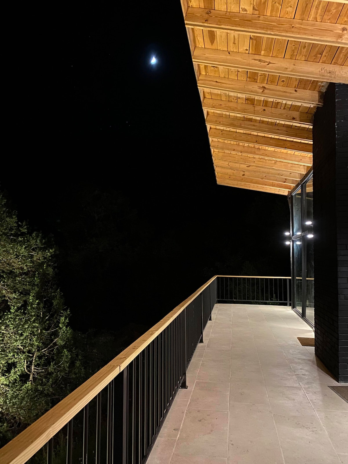 A spacious balcony is shown at night, illuminated softly by exterior lights. A wooden ceiling adds warmth, and the moonlight casts a gentle glow on the surrounding trees, creating a serene atmosphere. The railing frames the view of the natural surroundings.