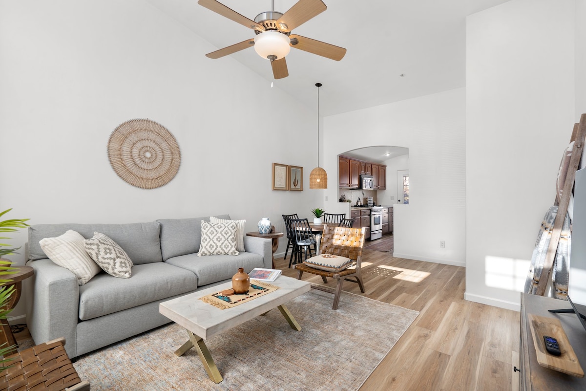A bright living room features a light gray sofa adorned with decorative pillows, complemented by a wooden coffee table. A ceiling fan hangs above, and natural light pours in through a window, illuminating the area. The space is connected to a dining area visible in the background.