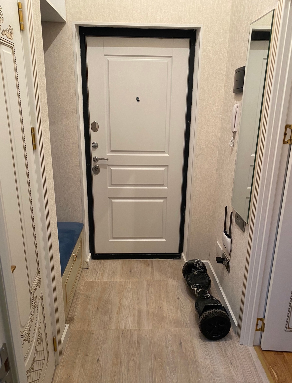 The entryway features a white door with a single doorknob and a keyhole, complemented by light-colored walls. A small bench is positioned to the left, and a personal transportation device is placed on the floor, adding convenience to the entrance.