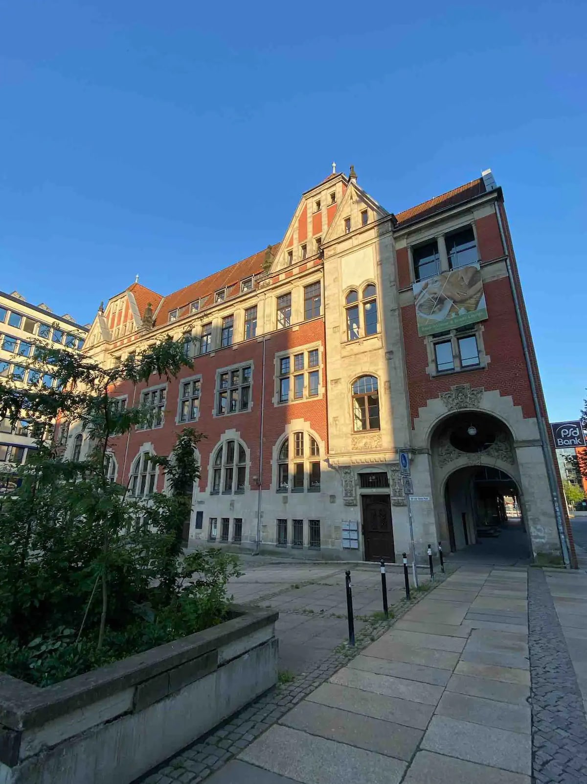 The historical building features a blend of brick and stone architecture, showcasing intricate details on the facade. Tall arched windows and decorative elements highlight its character. The entrance is accentuated by a prominent arch, with a pathway leading to a landscaped area in the foreground.