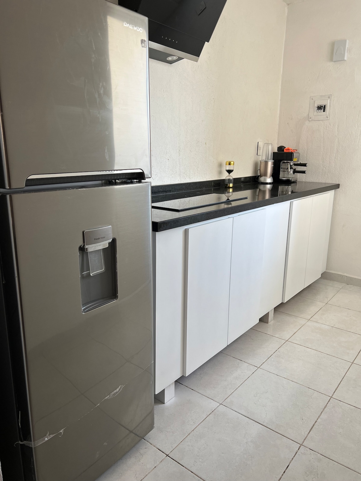 A modern kitchen setup is displayed, featuring a sleek stainless steel refrigerator next to a white cabinetry. A black countertop provides space for a coffee maker and other appliances, with tile flooring enhancing the clean and minimalist look.