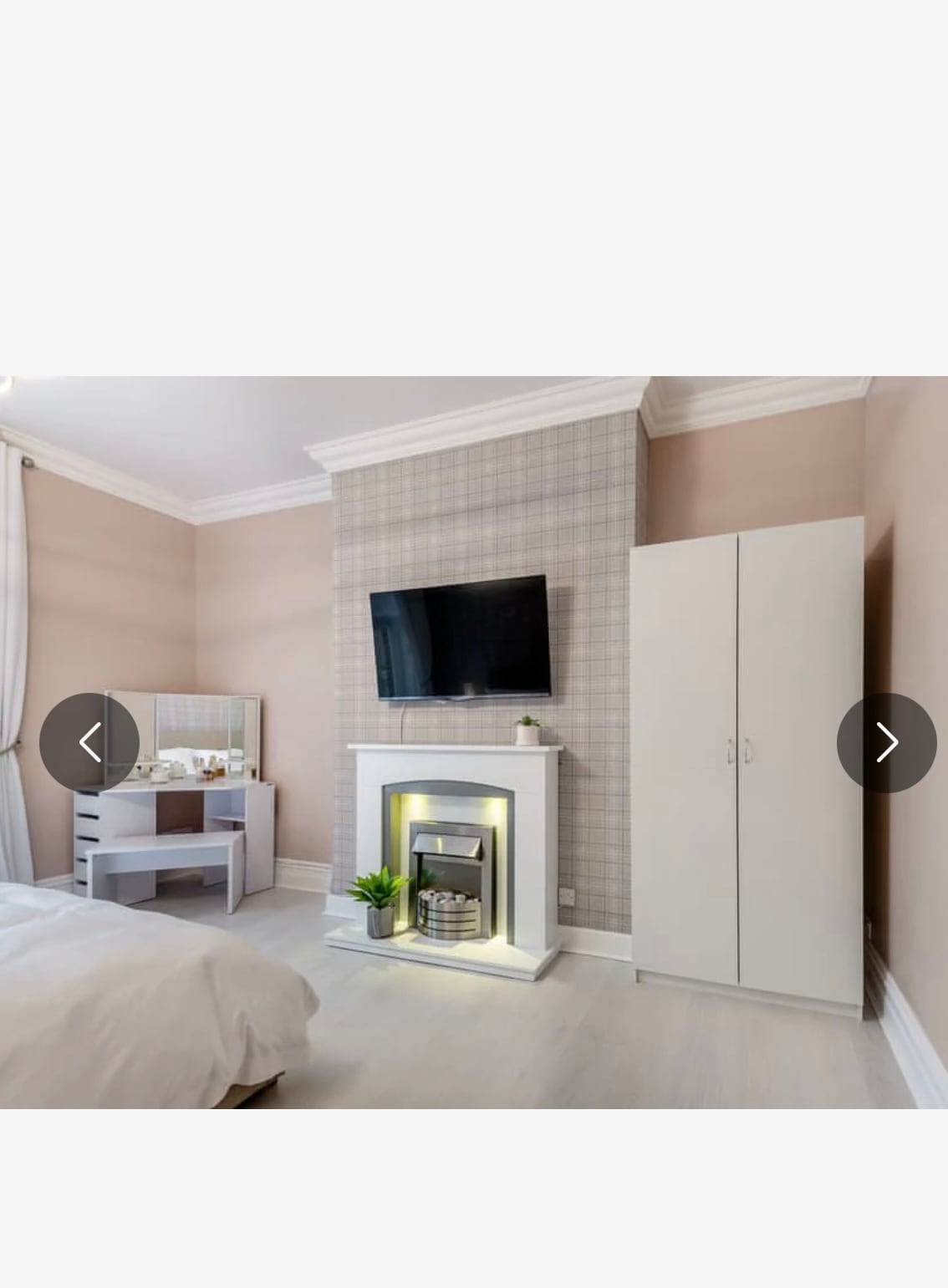 A softly lit bedroom features a stylish electric fireplace beneath a wall-mounted television. A light-toned wardrobe stands next to a desk with a mirror, offering a contemporary design. The neutral walls are complemented by light flooring, creating a calm and spacious atmosphere.