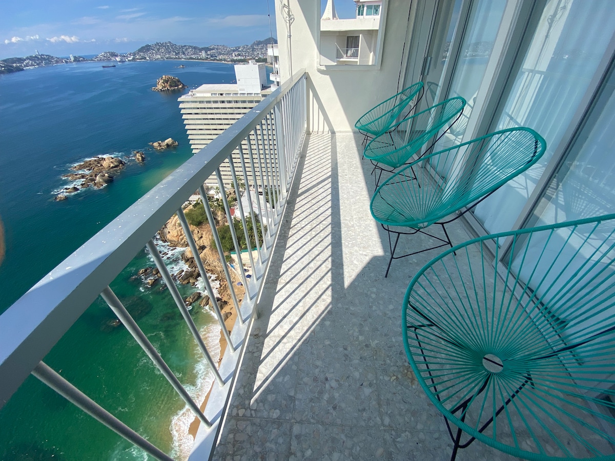 A balcony overlooking the sea features three teal chairs arranged on a light stone floor. The railing provides a safety barrier while allowing for unobstructed views of the rocky coastline and distant hills under a clear blue sky.