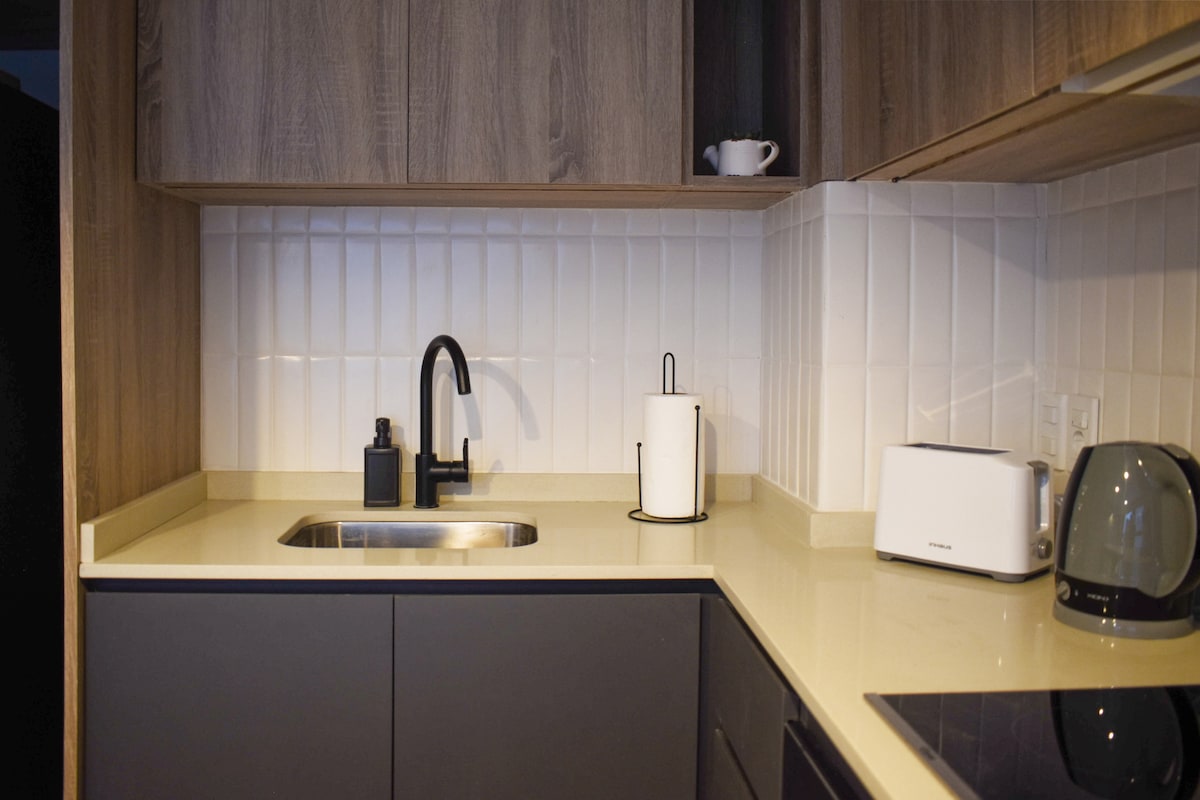 A modern kitchen area features wooden cabinetry complemented by a light-colored countertop. An elegant black faucet is positioned above the stainless steel sink. Small appliances such as a toaster and a kettle are arranged neatly on the surface, while a white dish holder adds a touch of simplicity.