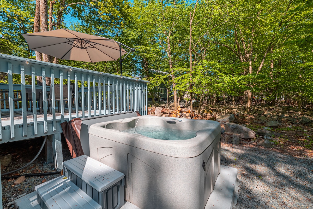 A hot tub is positioned on a wooden deck, surrounded by lush greenery. An umbrella provides shade, while steps lead up to the tub. The serene setting invites relaxation amidst nature.