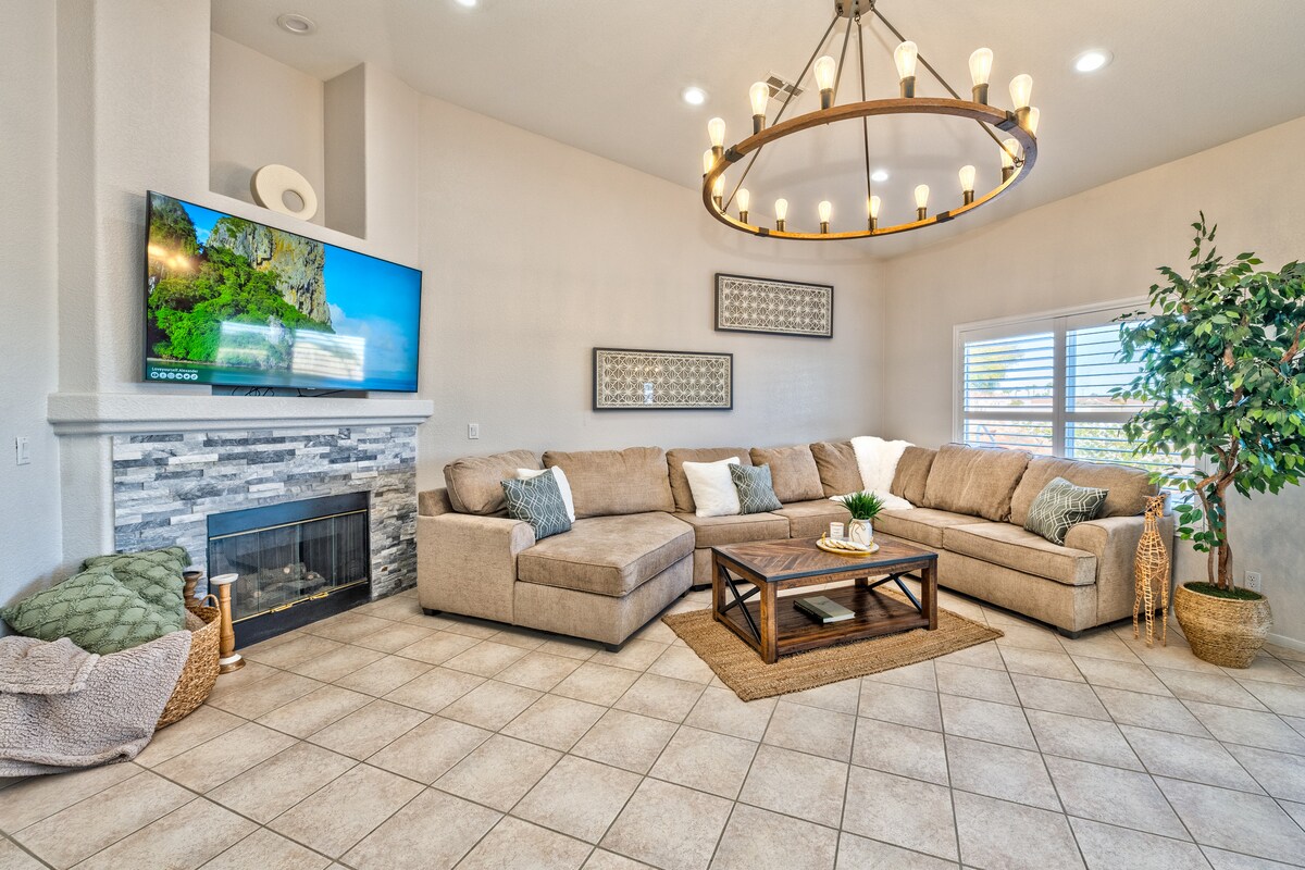 A cozy living area features a spacious sectional sofa arranged around a wooden coffee table. A large flat-screen TV hangs above a modern fireplace, while a unique chandelier illuminates the room. Large windows allow natural light to fill the space, complemented by indoor greenery.
