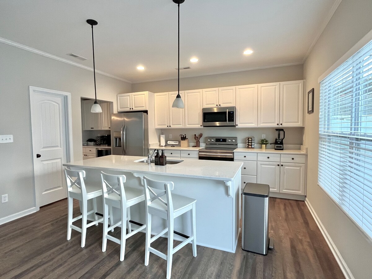 A modern kitchen is equipped with stainless steel appliances, including a refrigerator and oven. A central island with seating for three is complemented by white cabinetry. Natural light streams through the nearby window, illuminating the clean, minimalist design.