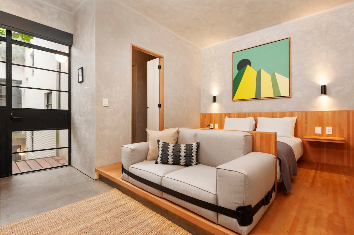 A bright room features a cozy seating area with a light-colored sofa adorned with decorative pillows. A spacious bed is positioned against a textured wall, complemented by bedside lighting. Large glass doors offer a view of the outdoors, enhancing the overall openness of the space.