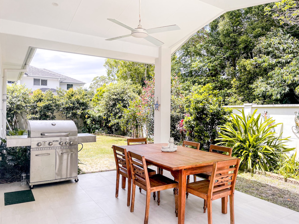 An outdoor dining area is shown featuring a wooden table surrounded by six matching chairs. A stainless steel grill is positioned nearby, and lush greenery is visible in the backdrop, complemented by colorful plants along the edges.