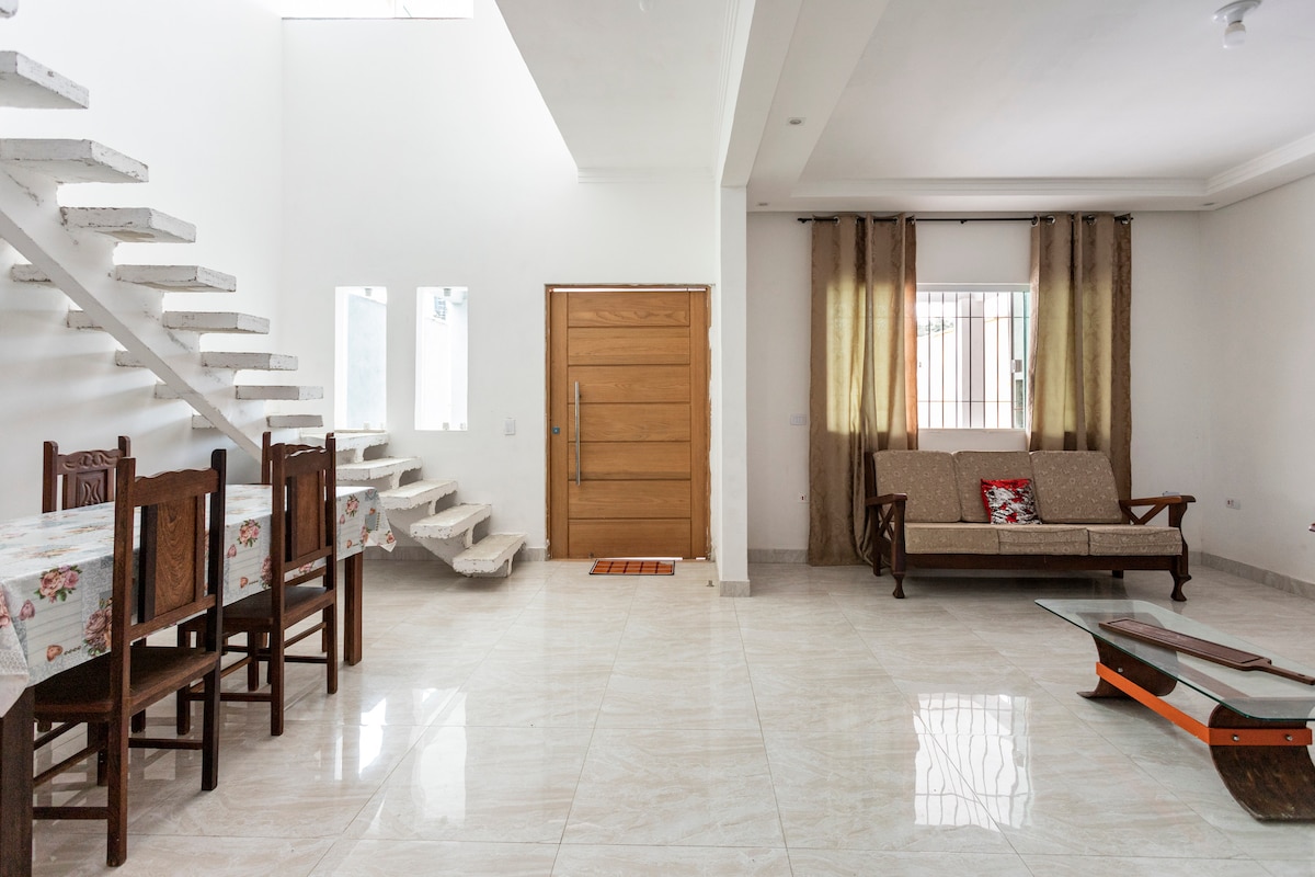 An open living space features a staircase leading to the upper floor, complemented by large windows allowing natural light. A dining table is positioned beside the entrance, while a cozy seating area with a sofa sits opposite, enhancing the home's spaciousness.