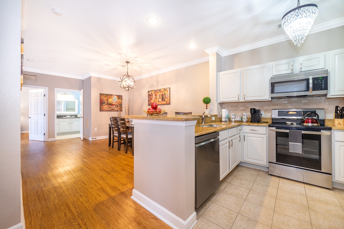 A modern kitchen is featured with elegant cabinetry, stainless steel appliances, and granite countertops. A dining table is positioned to the left, surrounded by several chairs. Natural light enters through a nearby doorway, enhancing the sleek tile and hardwood flooring throughout the space.