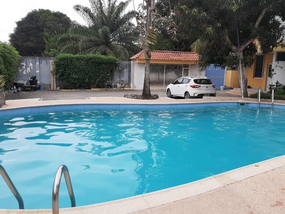 A clear swimming pool is framed by a textured stone deck, inviting relaxation. Lush palm trees and greenery surround the area, providing a sense of privacy. A light-colored vehicle is parked nearby, with a residential building visible in the background.
