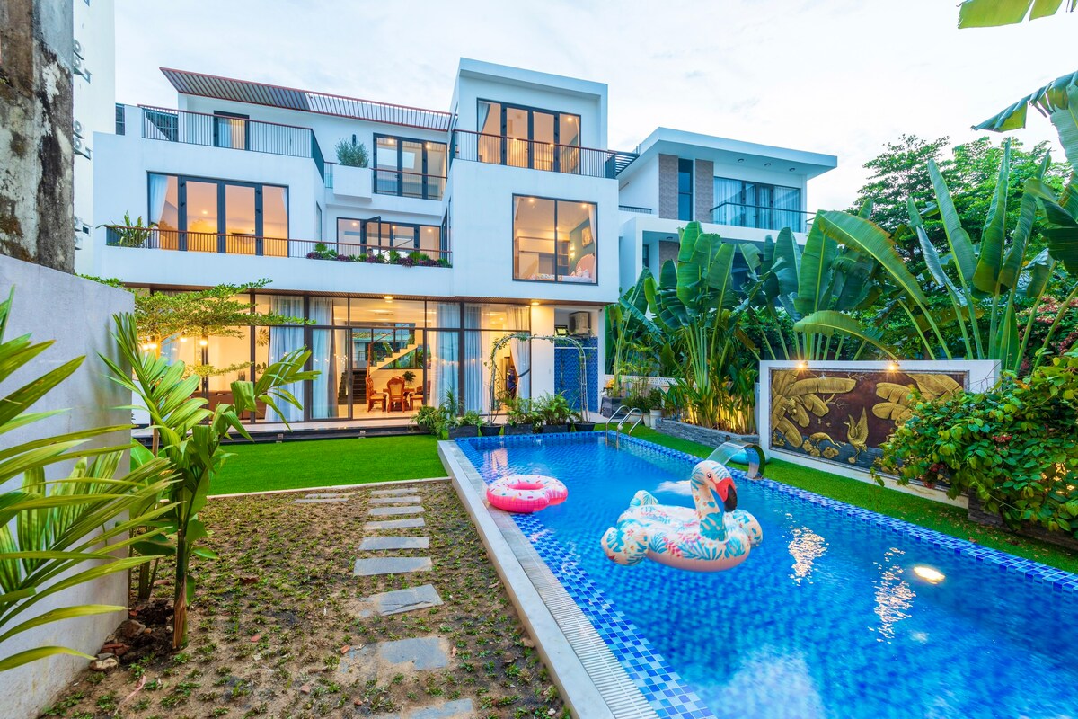 A modern villa is set against a vibrant garden, featuring a private swimming pool surrounded by lush greenery. Colorful pool floats are visible in the crystal-clear water, while large windows provide a view of the inviting living spaces within.