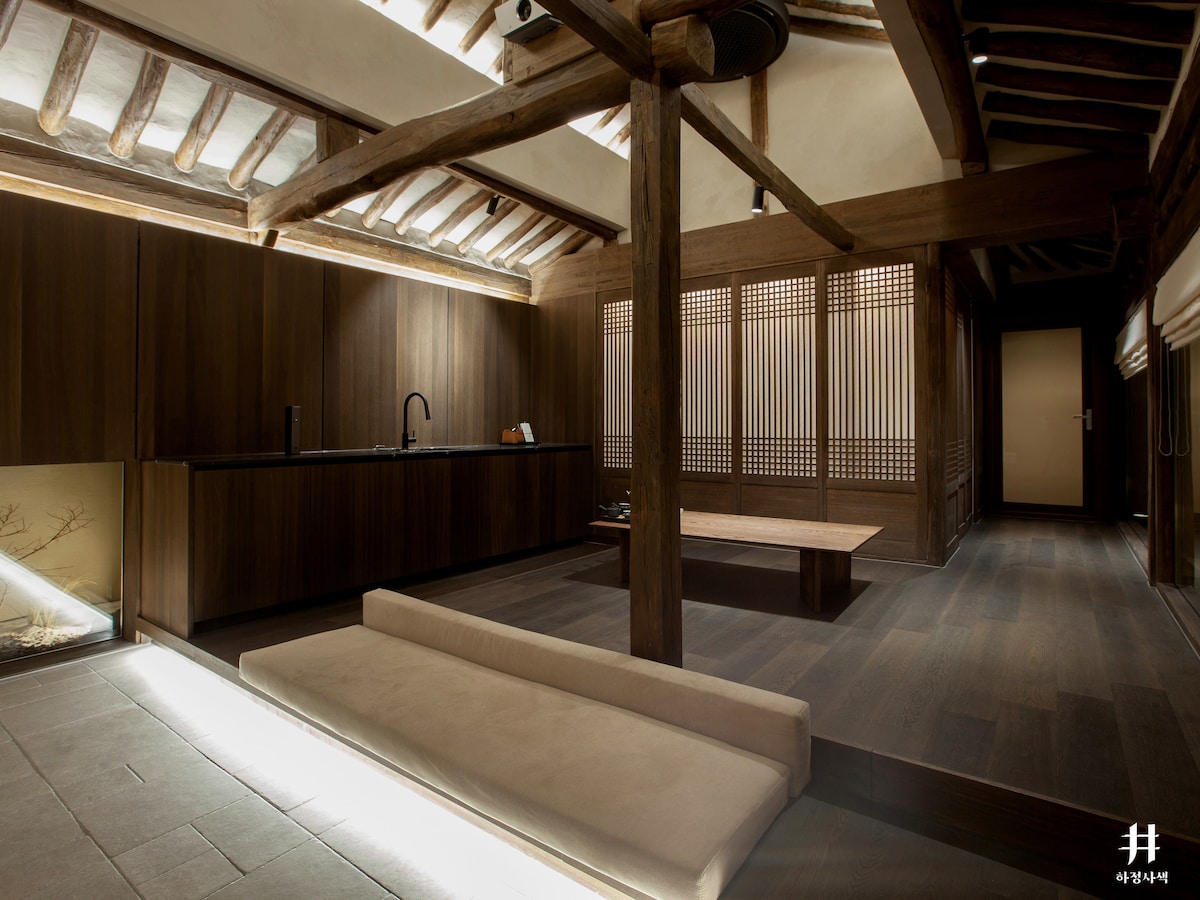 The interior showcases a blend of natural wood and stone elements, creating a calming environment. A low-seated, plush sofa is positioned on the expansive wooden floor, with a simple kitchenette featuring sleek black fixtures. Soft lighting highlights the structural beams overhead, enhancing the serene atmosphere.