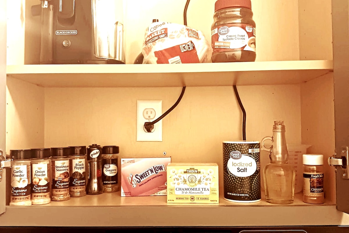 A well-organized kitchen shelf displays an assortment of spices, including onion powder and garlic pepper. Sweeteners and a box of chamomile tea are also visible, along with a container of iodized salt. A clear bottle is placed next to the spices, adding functionality to the space.